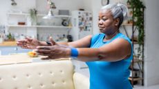 A woman exercises with a resistance band inside a house. She is standing with her arms out in front of her and a short looped resistance band around her wrists. Her hair is tied back and she is wearing a t-shirt. Behind her we see a couch, shelving and a decorated fridge. 