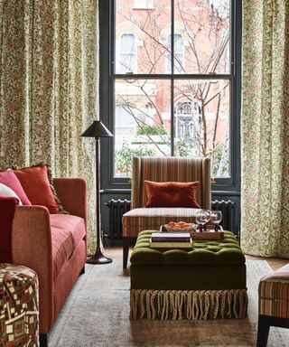 living room with grey window frames, botanical leafy drapes, coral couch, stripe armchair, green footstool, grey rug, floor lamp