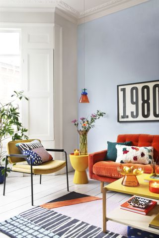 colorful living room with pale blue walls, orange couch, yellow chair, coffee table and side table, colored rug, bright patterned cushions, vase of flowers, artwork