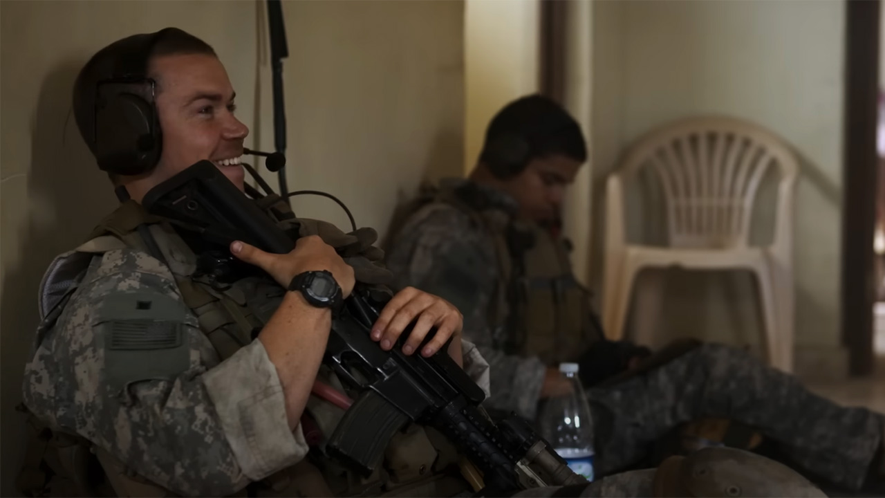 Will Poulter holding a gun and smiling amidst chaos in fatigues in Warfare.