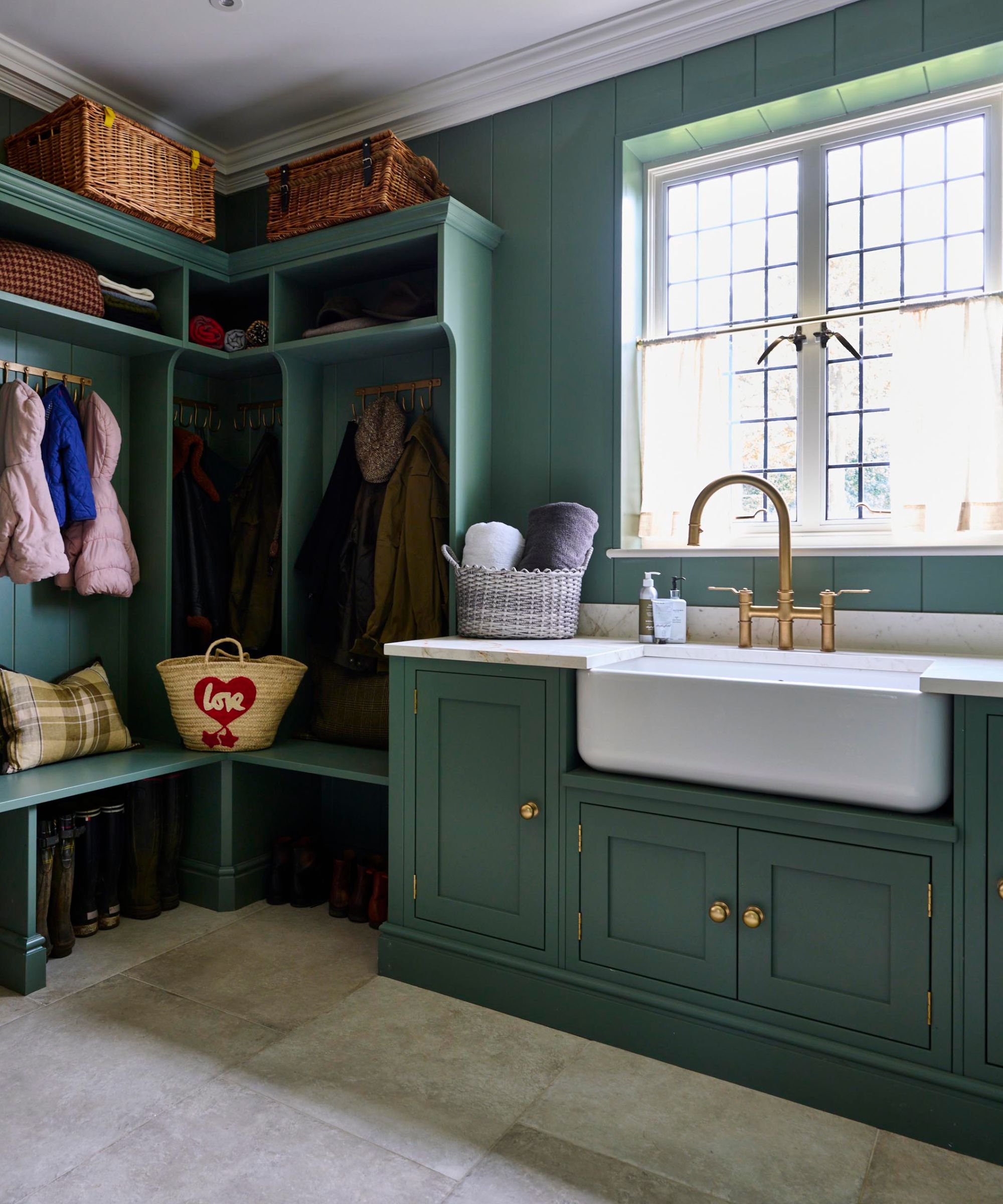 This multi-functional boot room combines smart storage, coat hooks, and a deep Belfast sink, all wrapped in a rich forest green scheme with classic brass hardware.