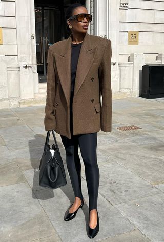 A woman wearing a brown blazer over a black t-shirt with black leggings, black pumps, a black handbag, and brown sunglasses.