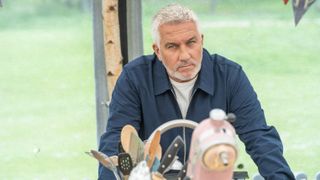 Paul Hollywood standing leaning against a table in the Bake Off tent wearing a dark blue shirt and looking stern