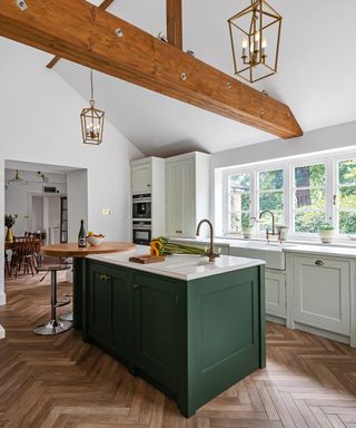 A bright kitchen with wooden beams on the ceiling, a dark green kitchen island, a large window, white cabinets, and dark wooden flooring