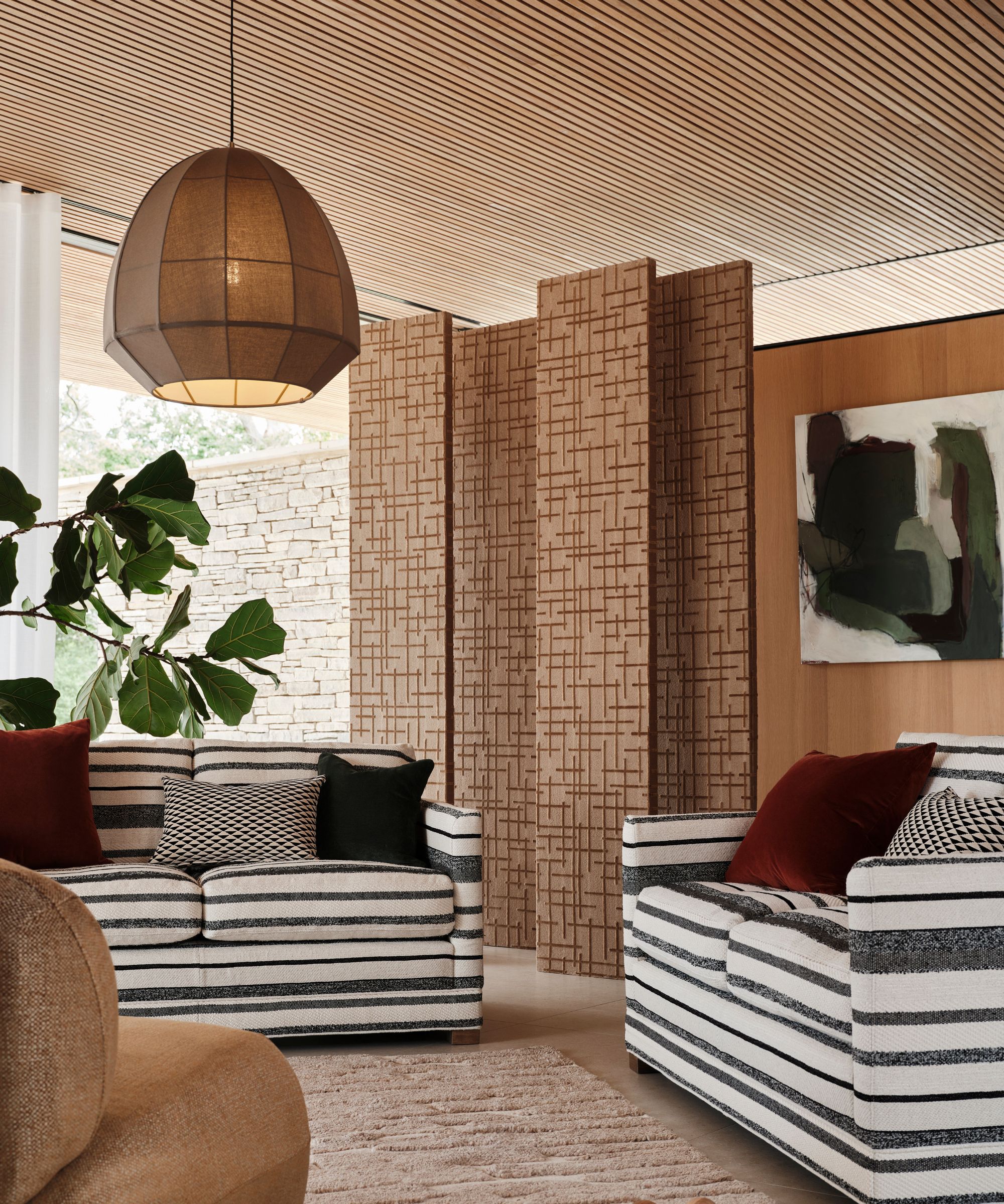Striped sofas with red velvet cushions face each other beneath a fabric pendant light and slatted screen.