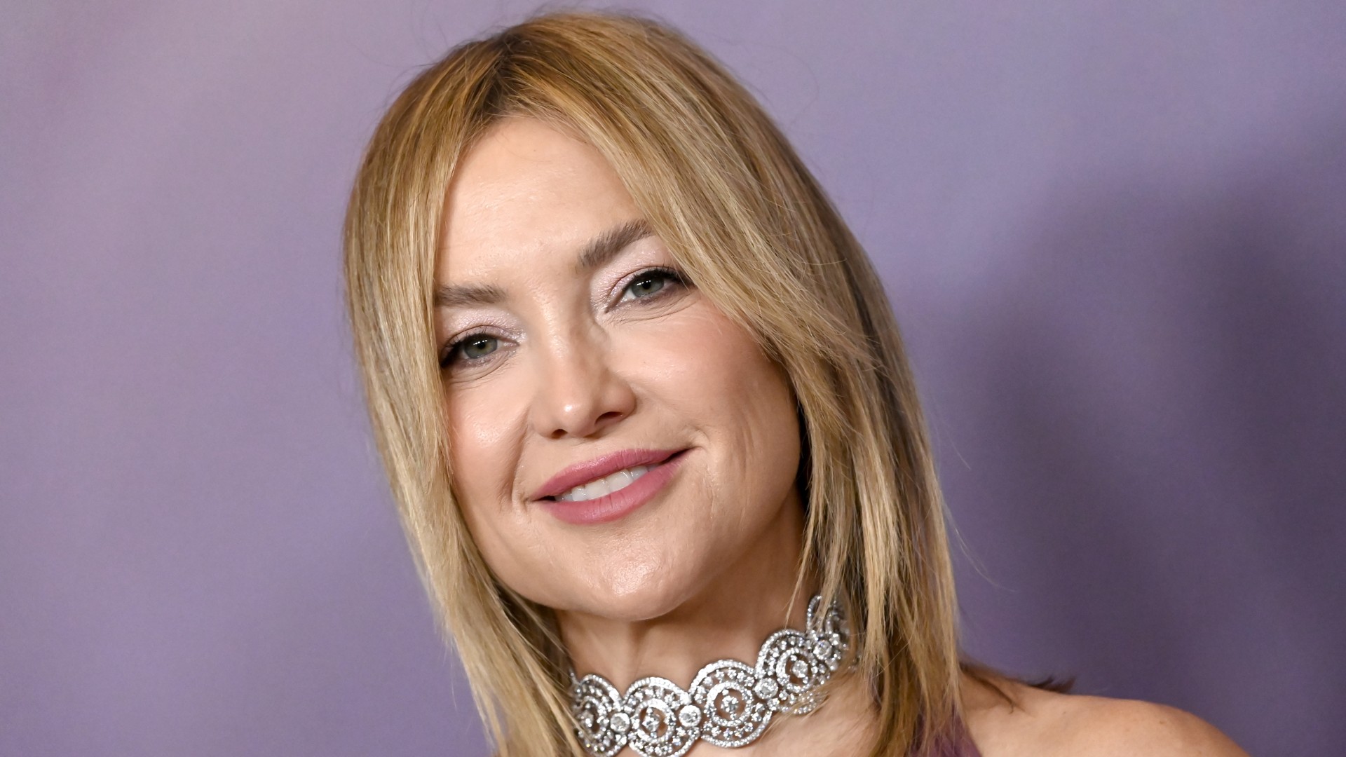 a white woman with short, blonde hair poses in front of a lavender backdrop