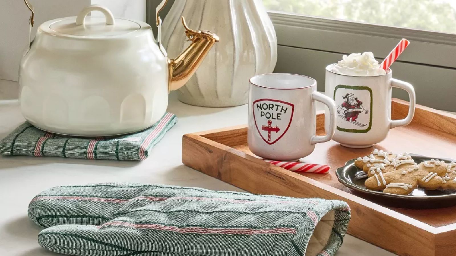 Green and red plaid oven mitt next to wooden tray with white hot cocoa mugs and gingerbread cookies