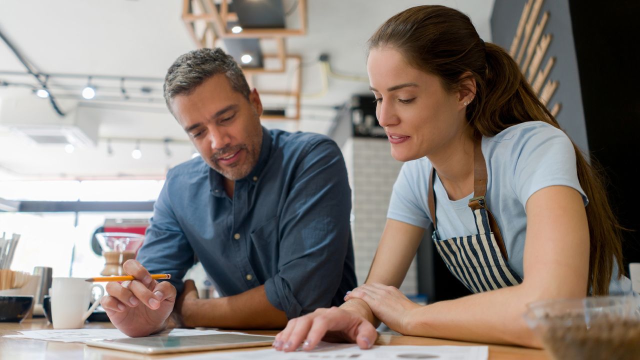 An adviser and coffee shop owner go over paperwork at the coffee shop counter.