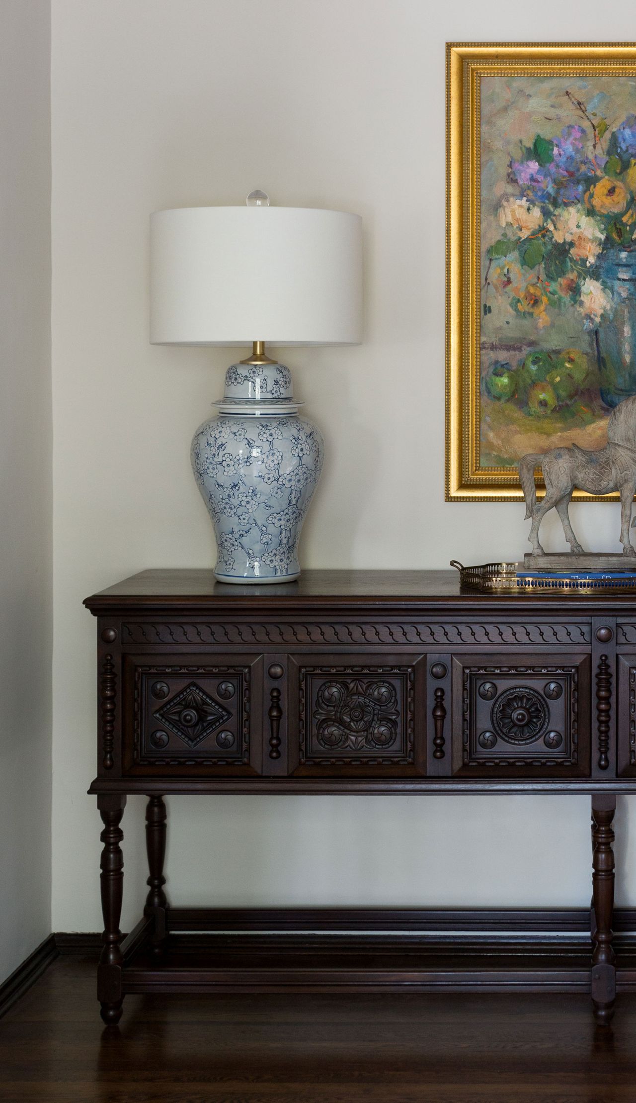 carved sideboard set against a white wall with gilt framed flower still life and traditional table lamp and dark wood flooring