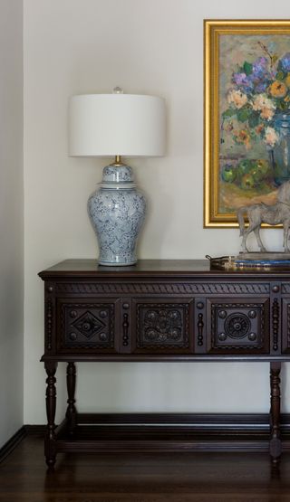 carved sideboard set against a white wall with gilt framed flower still life and traditional table lamp and dark wood flooring