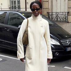 French influencer wearing white funnel-neck coat with brown back and black sunglasses, standing on Paris street.