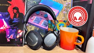 A pair of Bowers & Wilkins PX7 S3 wireless headphones stood upright on a table in front of some vinyl records and next to an orange mug