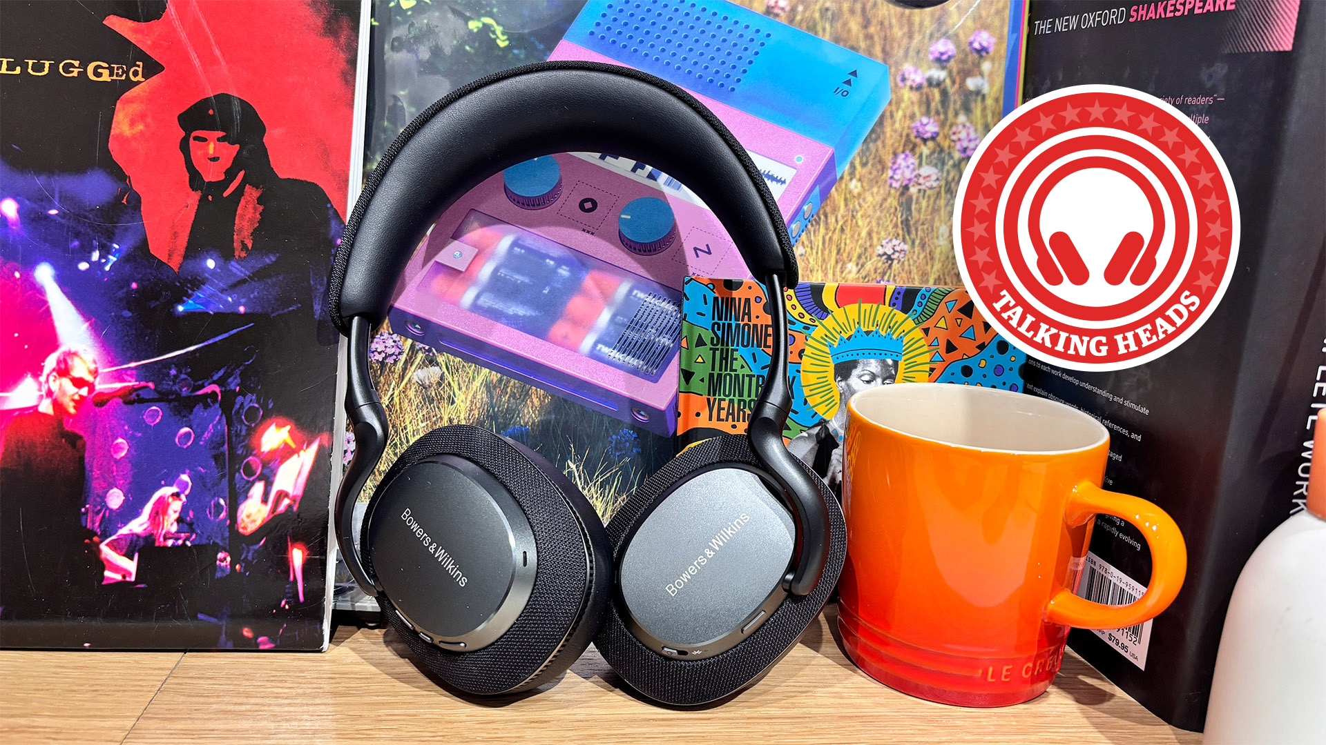A pair of Bowers & Wilkins PX7 S3 wireless headphones stood upright on a table in front of some vinyl records and next to an orange mug