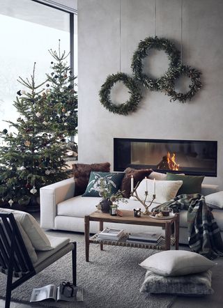 A modern Scandi Christmas-themed living room with trio of garlands on wall, cassette fireplace built into wall, and two Christmas trees