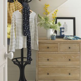 A wooden chest of drawers next to a coat stand holding clothing and scarves