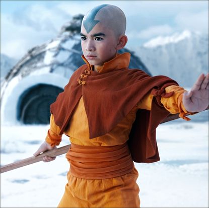 a bald-headed boy (Gordon Cormier as Aang) thrusts out his hand while holding a wooden staff and standing in front of an igloo, in episode 101 of Avatar: The Last Airbender