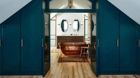 A view through bedroom double doors with blue floor to ceiling cabinetry either side, to a bathroom with a copper bath, wood stool by its side and double vanity centralised on the back wall.