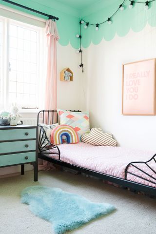 Kid's bedroom with white walls, green scalloped paint effect on ceiling and top fifth of wall, black metal bed frame, pink spotty bedding and green and black upcycled chest of drawers