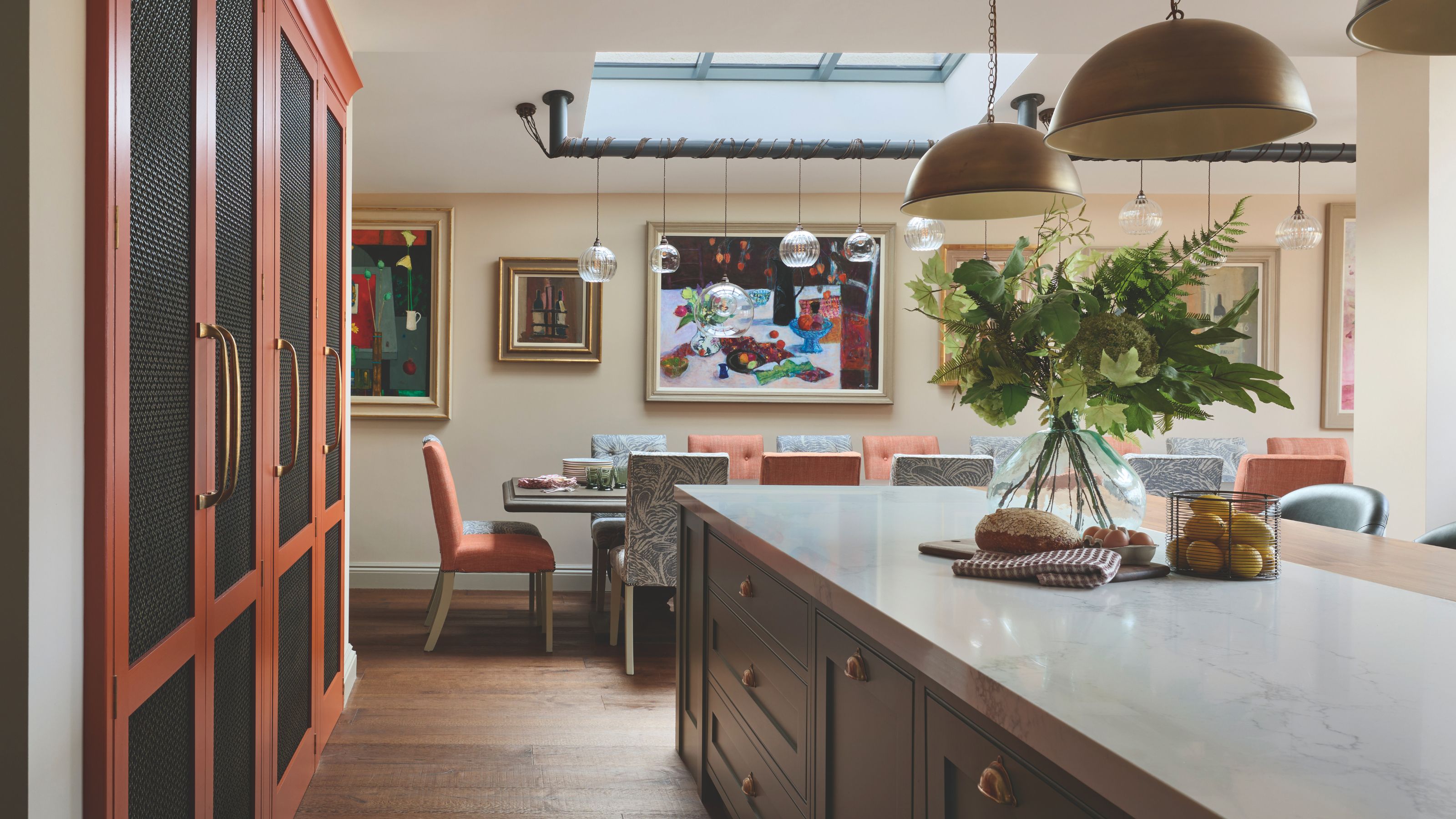 A vibrant, contemporary open-plan kitchen and dining room. In the foreground, a white marble kitchen island sits next to tall, rust-orange cabinets with dark mesh panels. The background features a long dining table with mismatched orange and patterned chairs, set under a skylight and eclectic pendant lighting, with colorful abstract artwork on the walls.