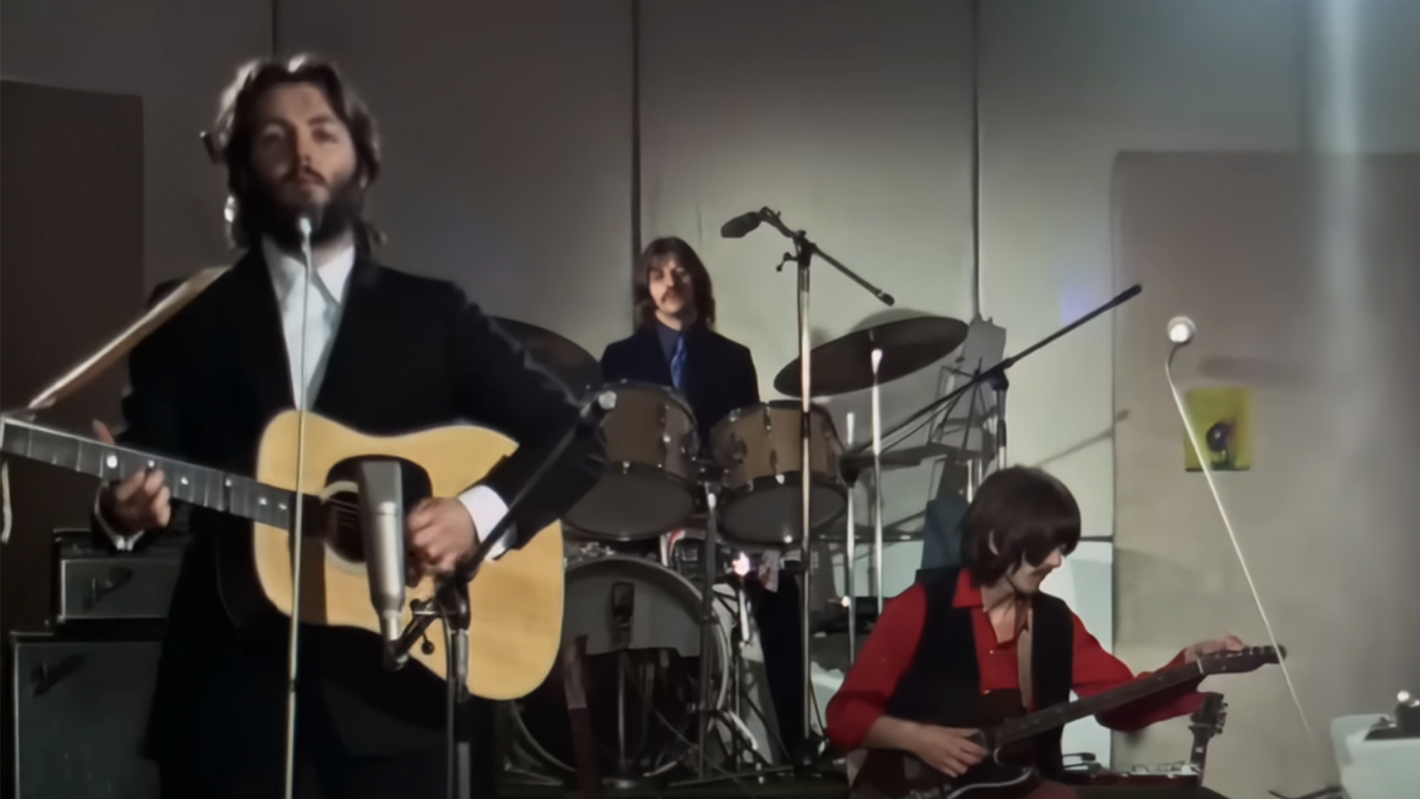 Paul McCartney playing an acoustic guitar in front of Ringo Star on drums and George Harrison playing guitar