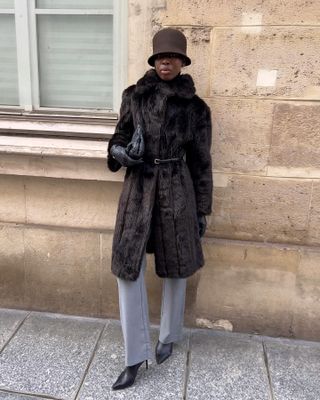 Sylvie wears a brown cloche hat, brown fur coat, gray jeans, and black boots.