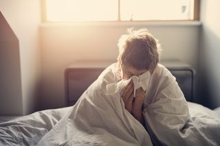 A boy in a bed with a duvet wrapped around his shoulders blowing his nose into a tissue