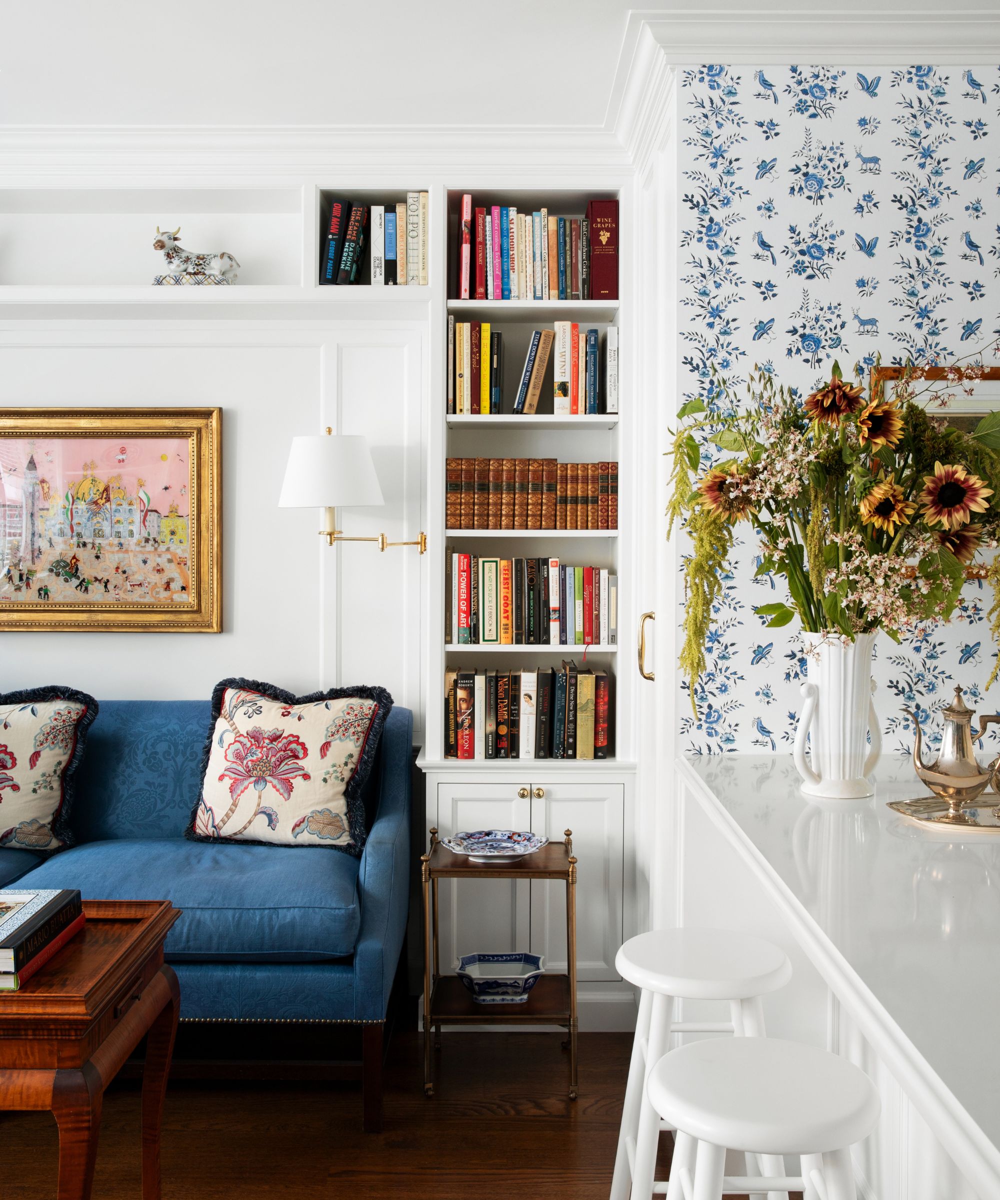 The corner of an adjoining small living room and kitchen with blue and white patterned wallpaper, built-in bookshelves, and a blue sofa
