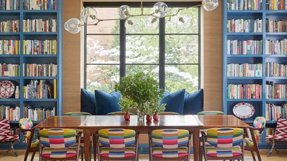 Mid-century modern dining room ideas are so chic. Here is a blue mid-century modern dining room with bookcases around the black grid window with a blue window seat underneath, and a teak wooden rectangular table with colorful chairs around it and a gold chandelier above it