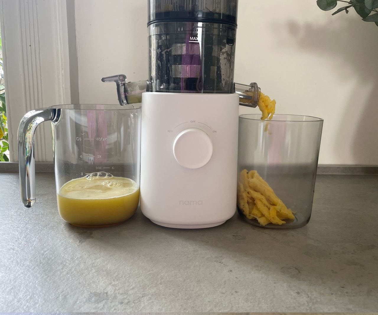 A close up of the base of a juicer, yellow fruit debris is funnelled into a beak on the right hand side, as fresh juice is poured into a container on the left.