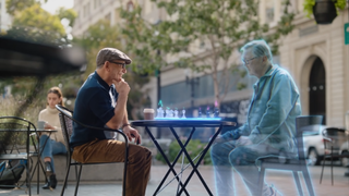 A man sitting at a table in public playing chess with an opponent in mixed-reality through Meta smart glasses. His friend is depicted as a hologram, as is the chessboard they're playing on.