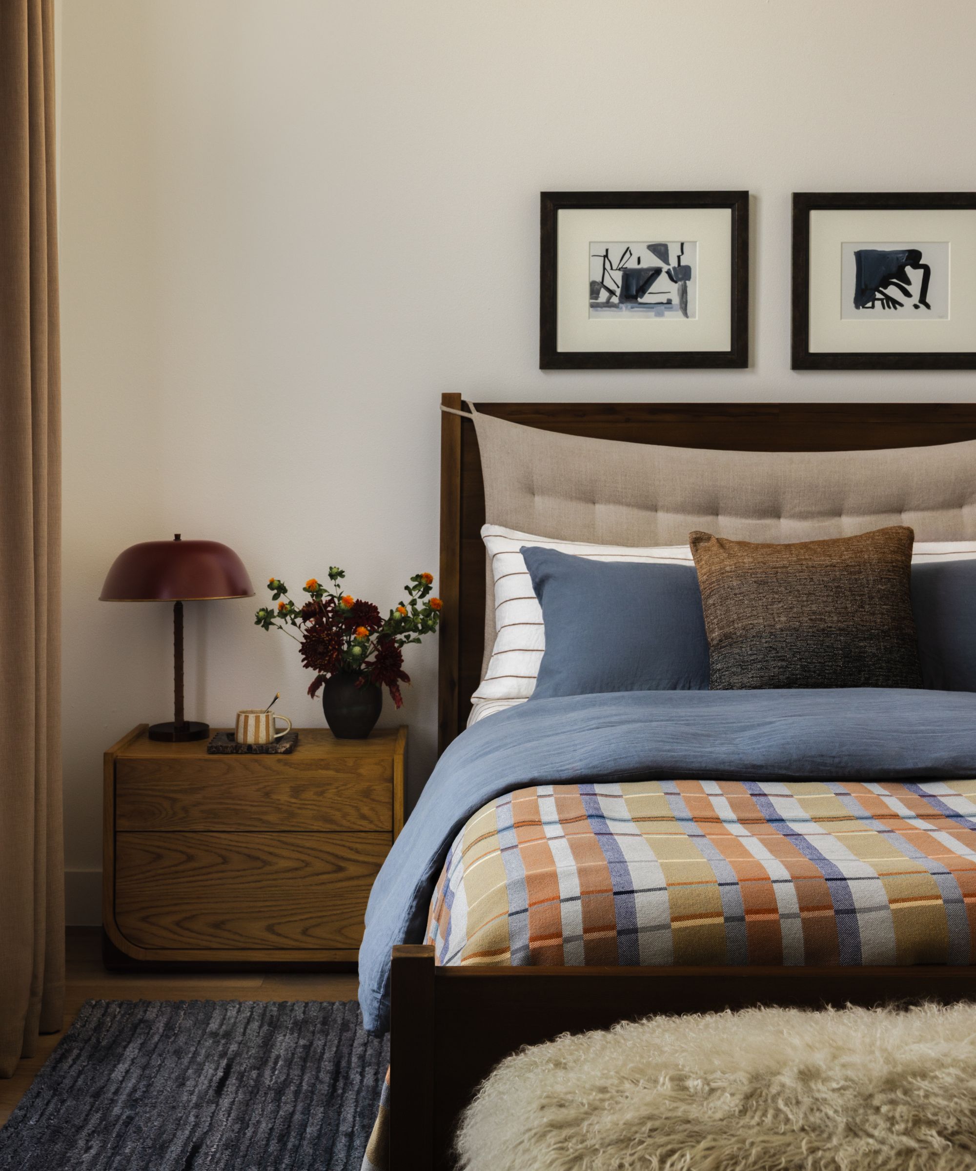 a neutral bedroom with blue and orange rust tones with a blue rug, wooden bed, plaid throw and oak nightstand
