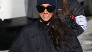 Meghan Markle wearing a black puffer jacket, with black sunglasses and a navy warm hat