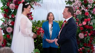 Billy and Honey getting married, Billy is in a navy suit and Honey is in a white bridal gown and there are red and pink flowers behind them