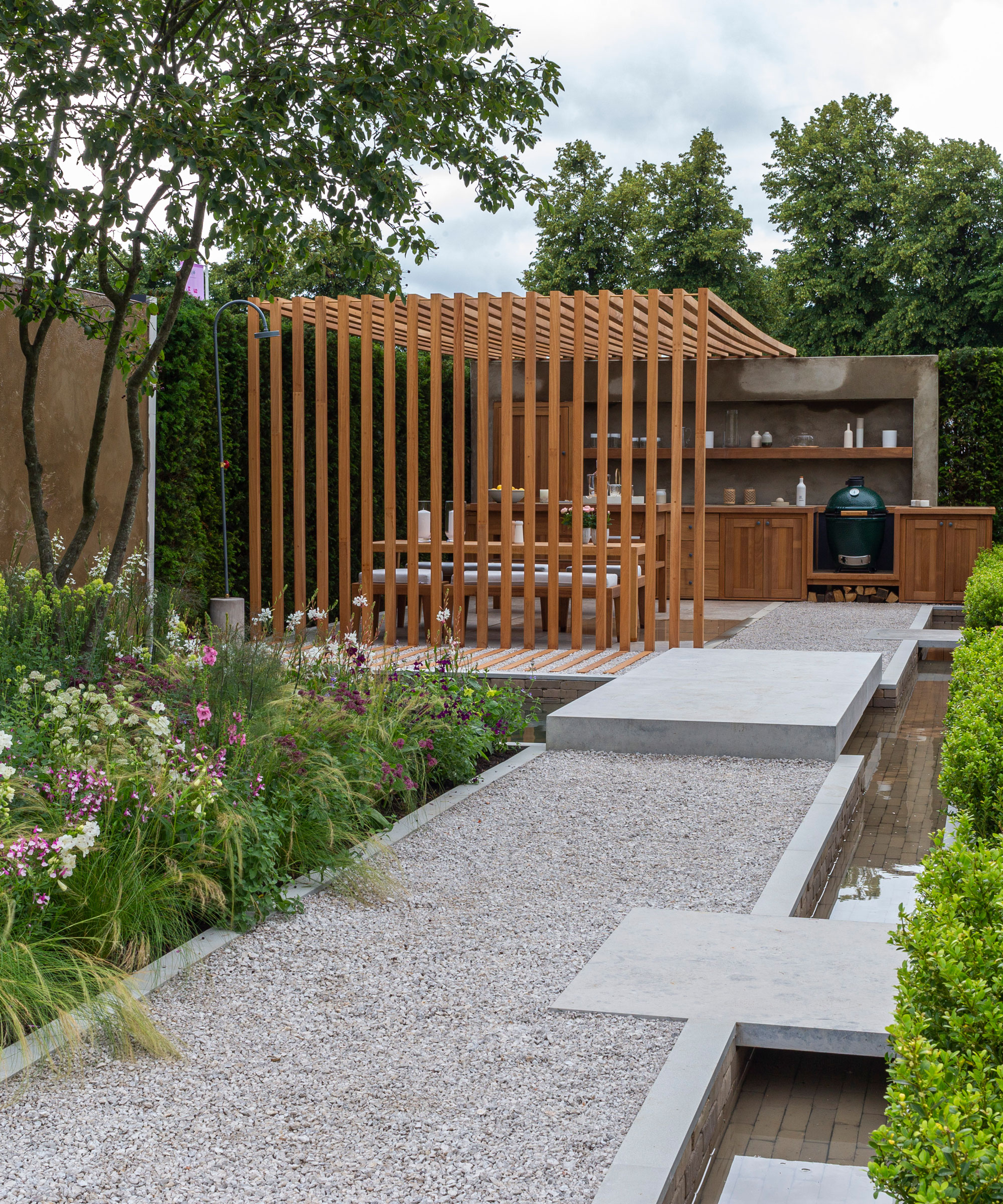 garden design with outdoor kitchen and bbq area, flowerbeds, raised gravel path, water feature and trees
