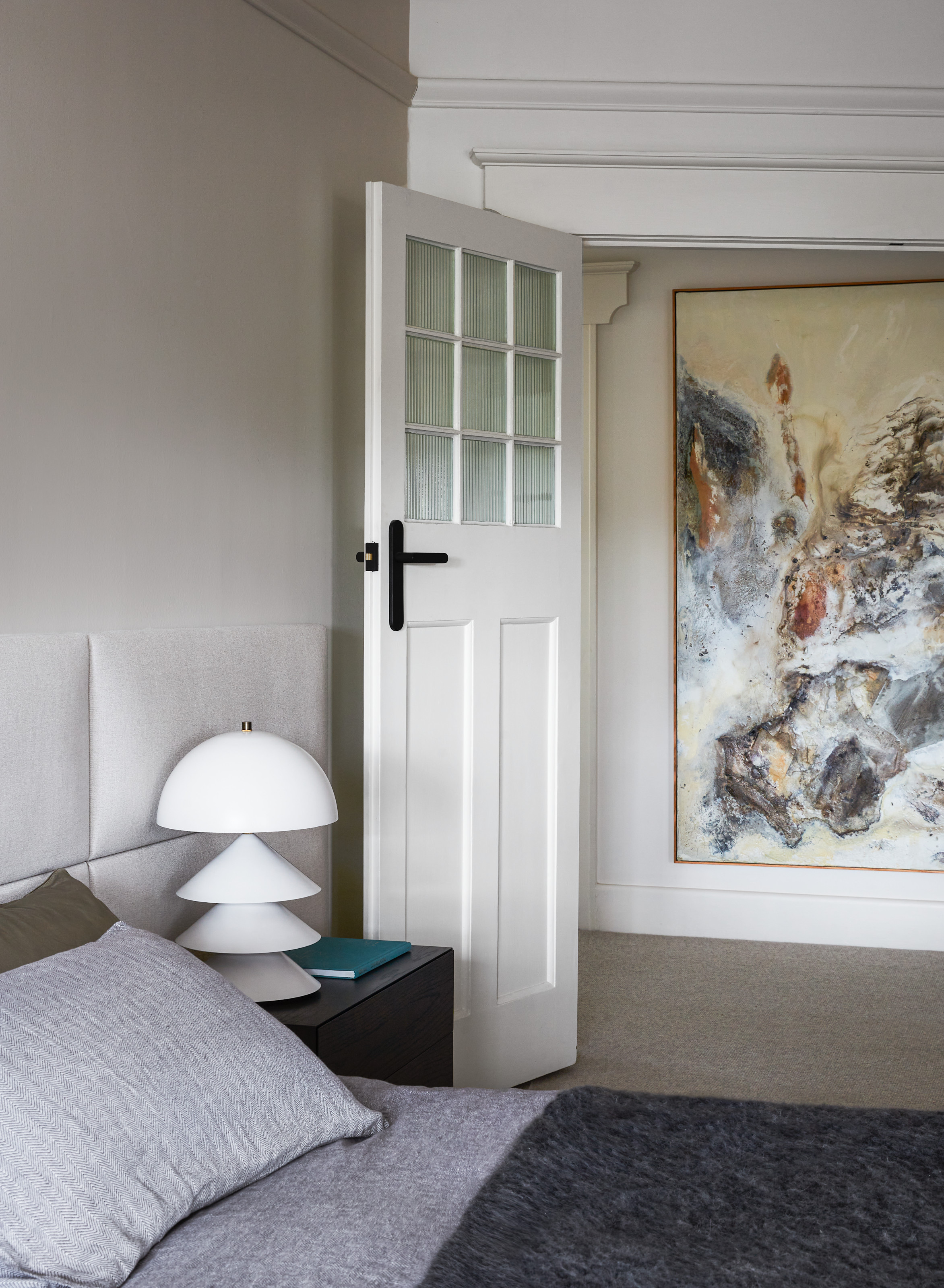 Bedroom with grey and white walls, pale grey extended headboard, grey bed linen and black side table with white stacked lamp