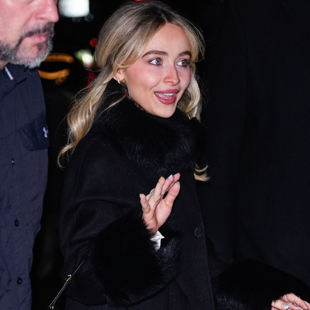 Sabrina Carpenter wears white satin platform heels with a longline black coat and a black leather handbag.