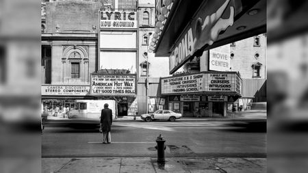 A man in a long coat stands alone at a street corner beside a fire hydrant, facing a row of cinema marquees advertising old films.