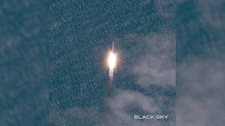 a white rocket launches above a plume of fire, with the sea visible behind it