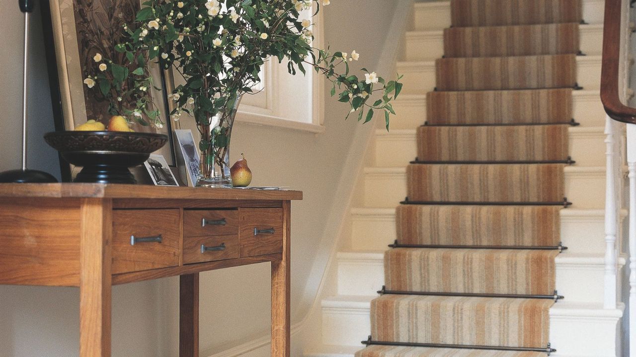 Staircase with carpet runner behind side table with fruit bowl, flower bouquet with vase, and portrait framed picture.