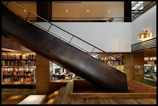 Interior view of Tsutaya Books featuring wood flooring, a dark coloured hammered steel staircase, white walls, pillars and shelving units filled with books