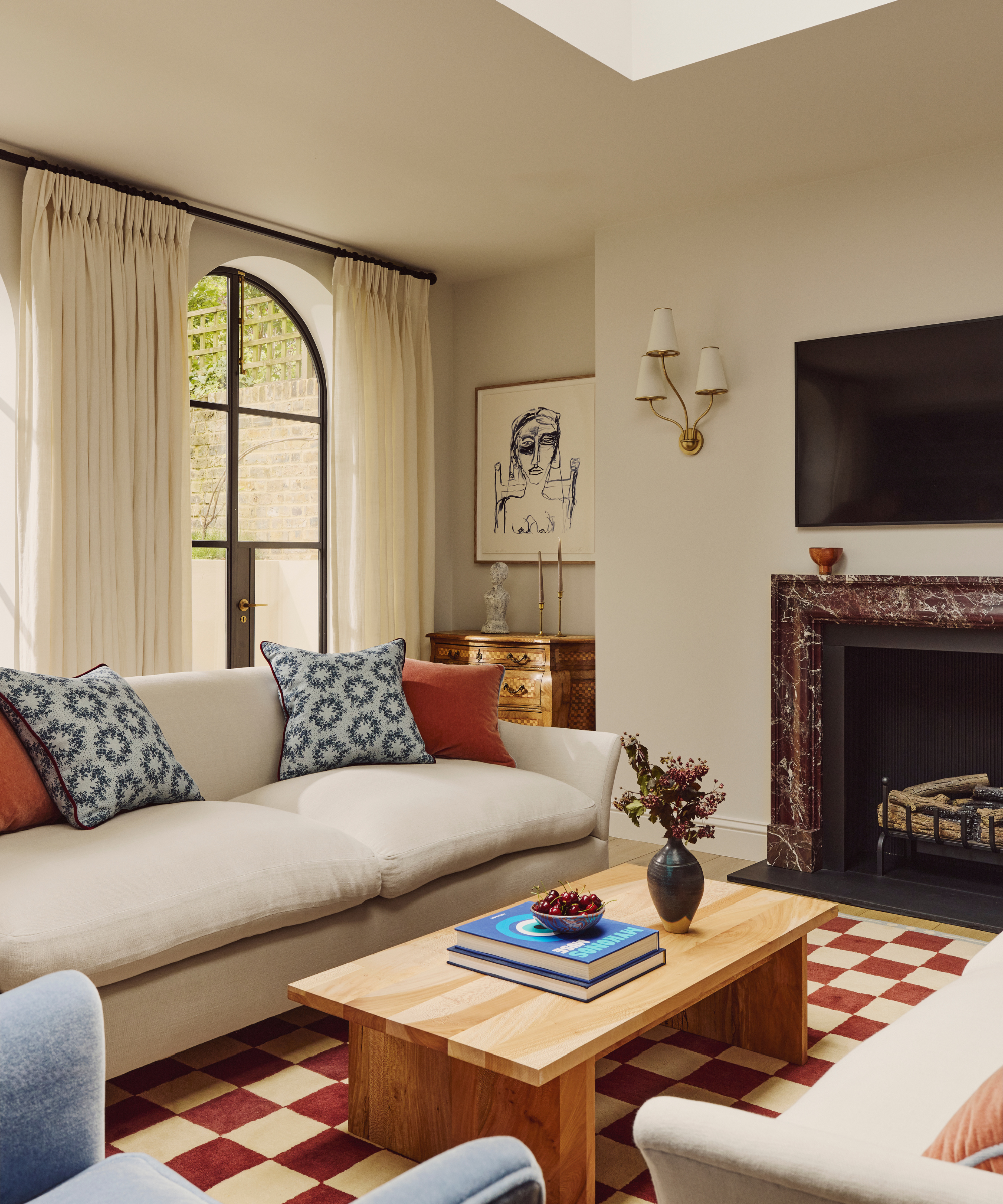 a living room with a checker-printed rug, a wooden coffee table, a cream-colored couch, decorative throw pillows, and tall windows with long white curtains hanging in front