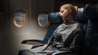 A woman asleep on a plane with a Trtl pillow and travel blanket