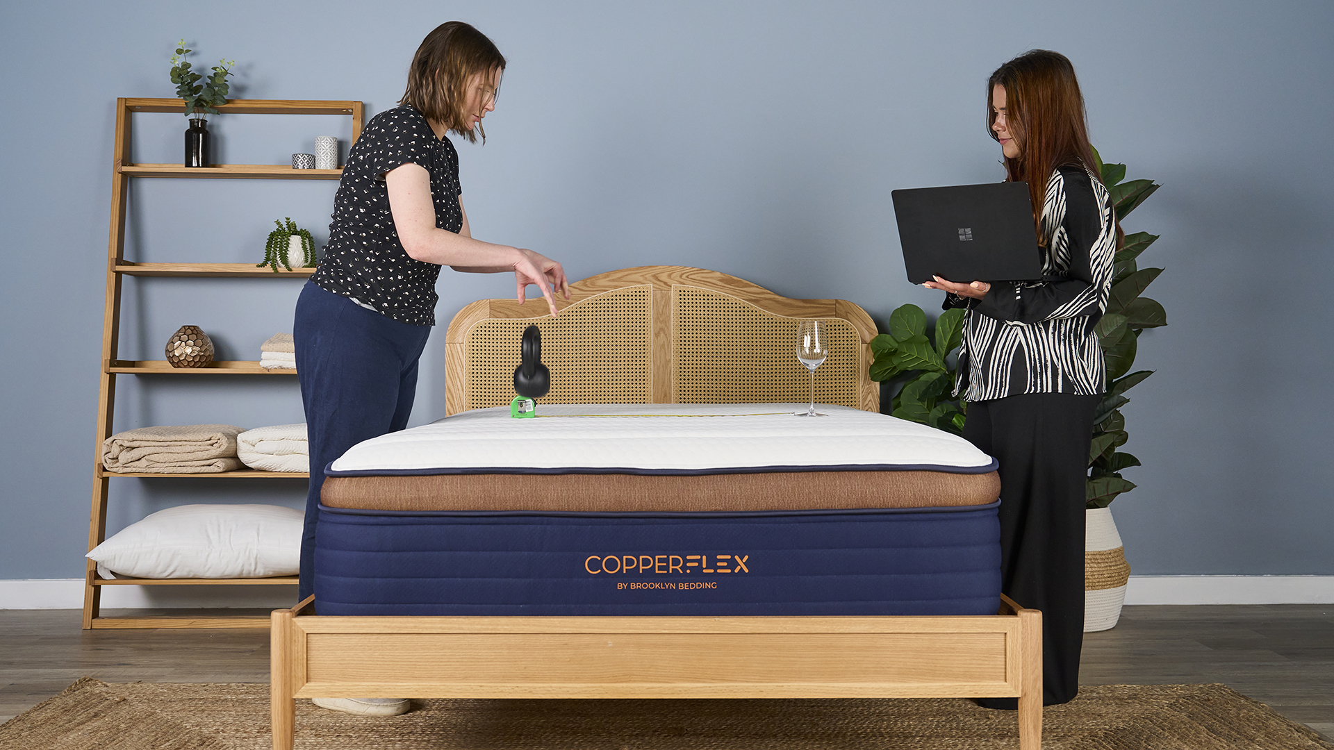 Two women carrying out a motion isolation test on the Brooklyn Bedding CopperFlex mattress in sleep studio. They place a wine glass on one side and drop a kettle bell on another.