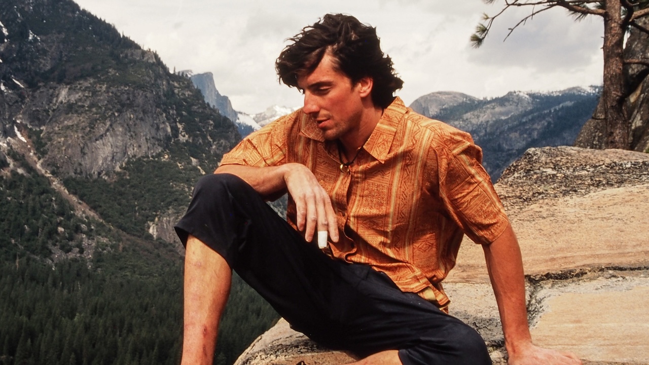 डोंगराच्या पार्श्वभूमीसमोर केशरी शर्ट आणि काळ्या पॅन्टमध्ये बसलेला डीन पॉटर