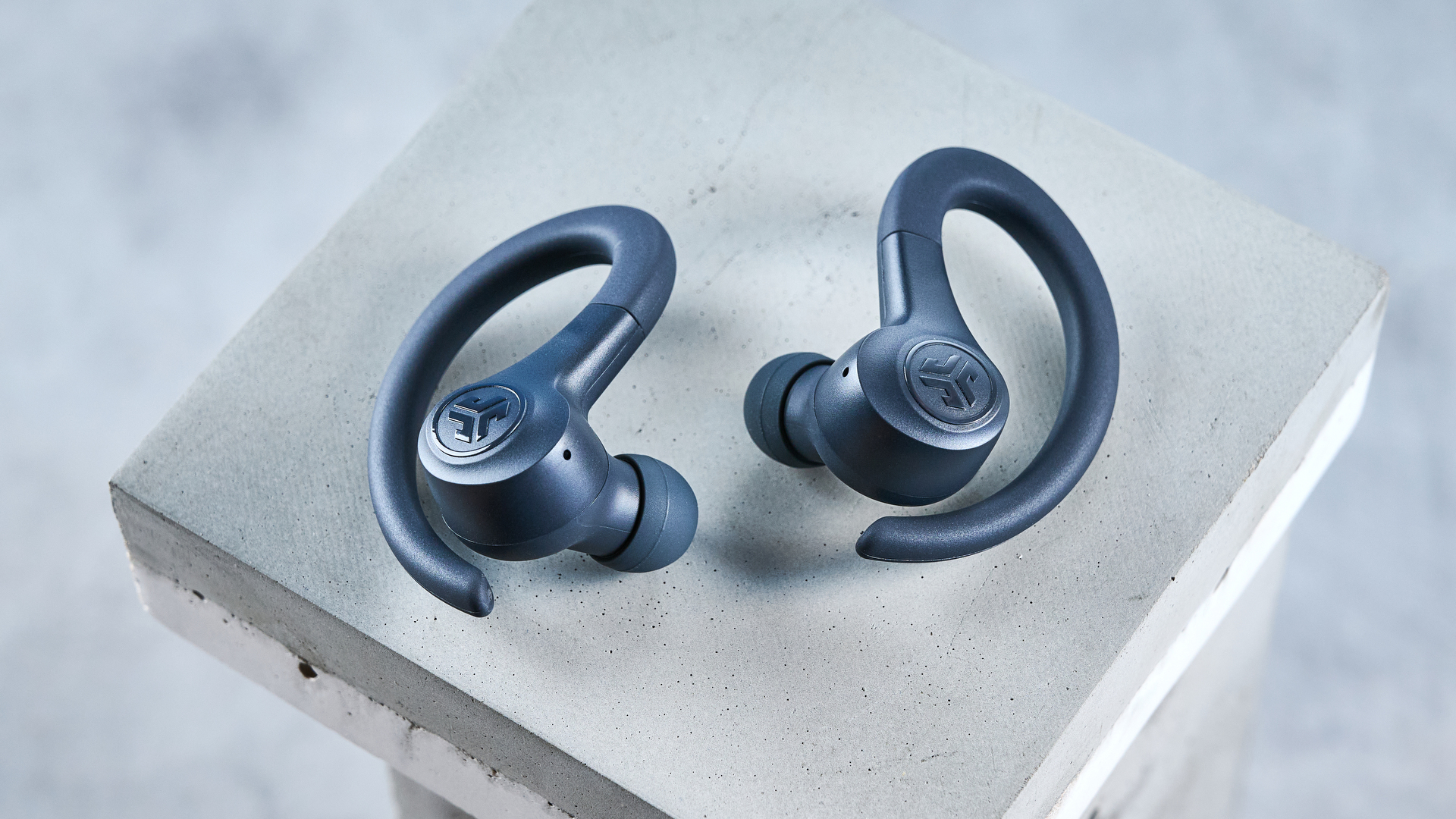 the jlab go sport+ earbuds photographed against the blue tom's guide background, showing charging case and wearing style