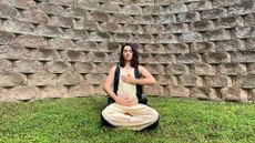 woman sat on some grass, holding one hand to her chest and the other to her belly, facing the camera with a stone wall behind her