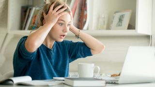 A woman sitting at a table with various objects on it, including a MacBook, a mug, a book, an opened notebook, and holding her head with her hands as if in frustration