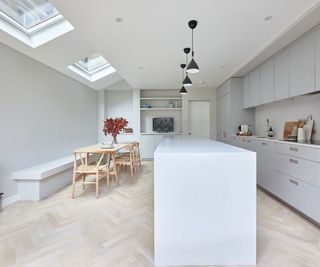 white and pale grey kitchen with long central island, light herringbone laid flooring, pendant lights and built in seating to one wall in front of dining table, with skylights above