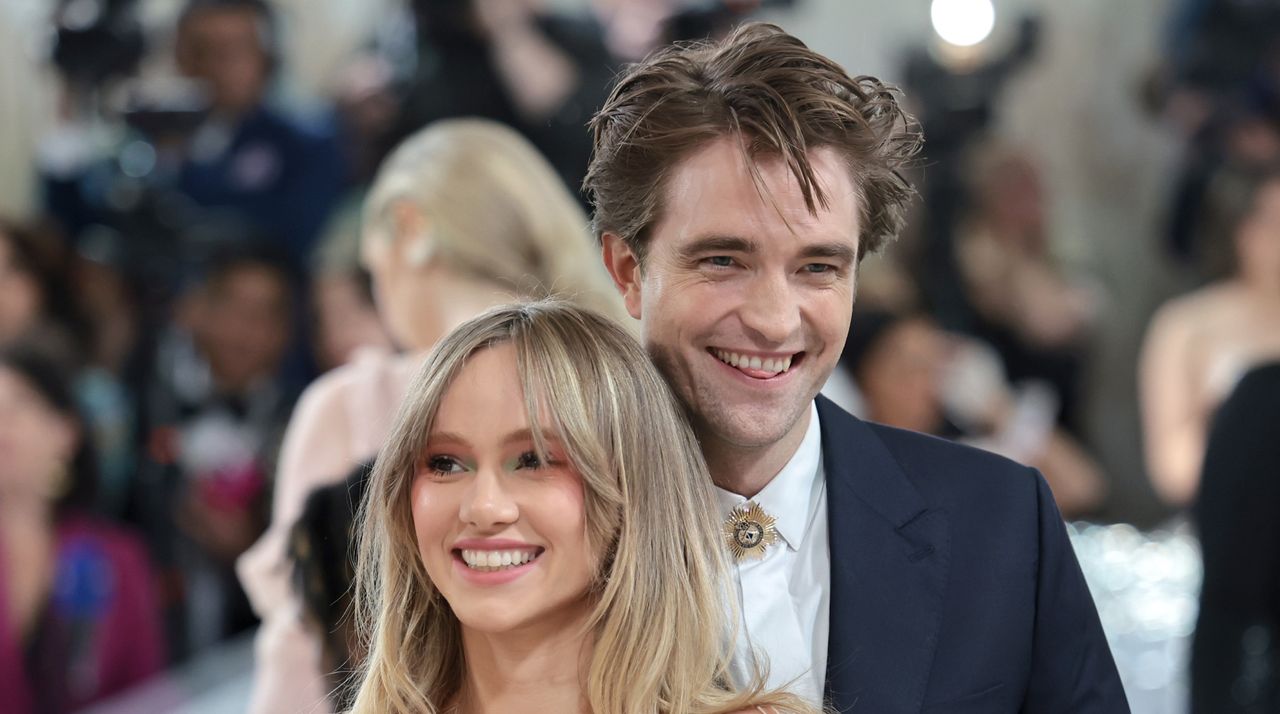 Robert Pattinson laughs and sticks his tongue out while wearing a white shirt and black suit on the Met Gala red carpet, and partner Suki Waterhouse smiles with long wavy golden blonde hair