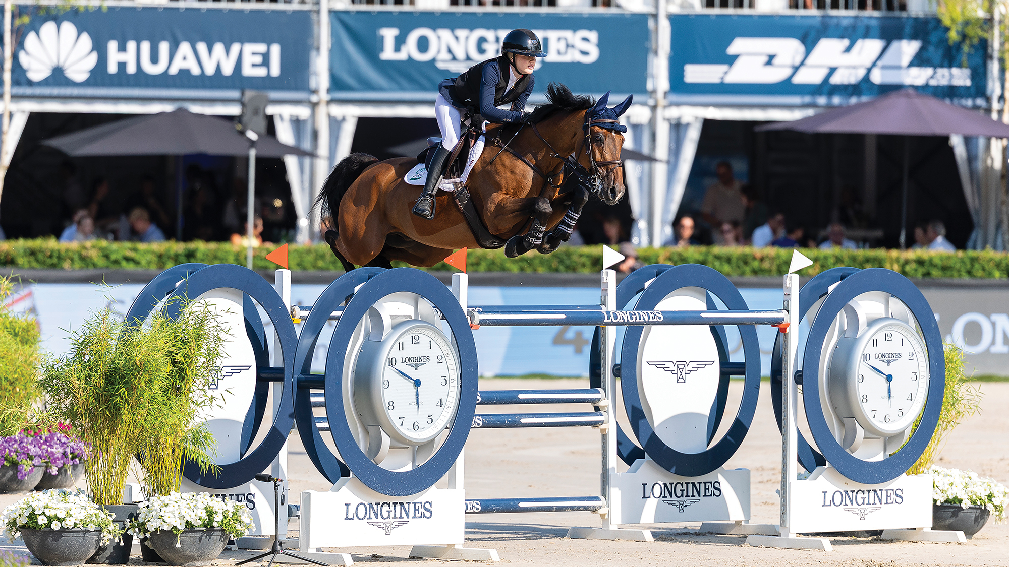British female showjumper Sienna Charles clears a huge blue and white Longines-branded oxer riding a bay horse at the LLN in Rotterdam